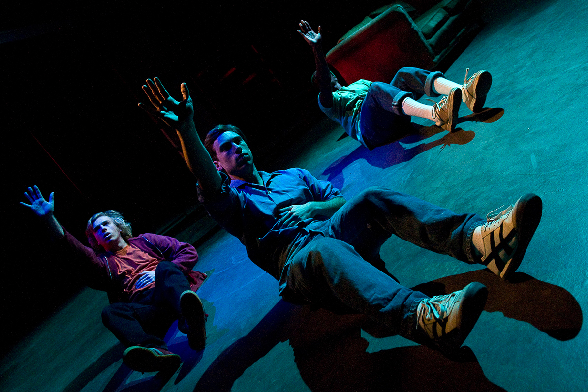 "Deviations" by Joe Goode Three performers sit on stage with their legs stretched out in front of them, their rights arms raised above their heads, and their left arms over their stomachs. They are illuminated with red, green, and blue lights.