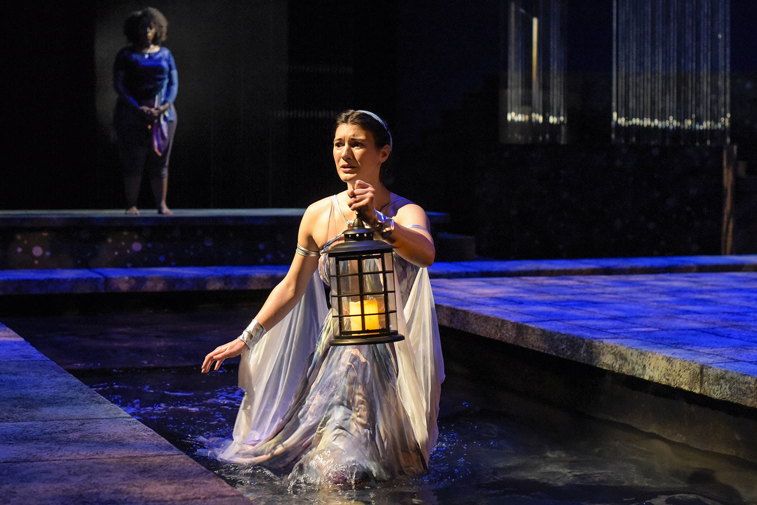 An actor wearing a lilac dress and holding a candlelit lantern wades through a pool of water on stage.