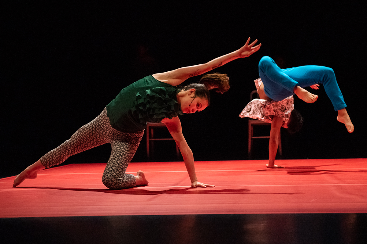 Two dancers perform on a stage with a red vinyl floor. The dancer in the foreground kneels on one knee while pointing their other leg at a 45 degree angle. The dancer in the background is balanced upside down on one hand while mid-flip.