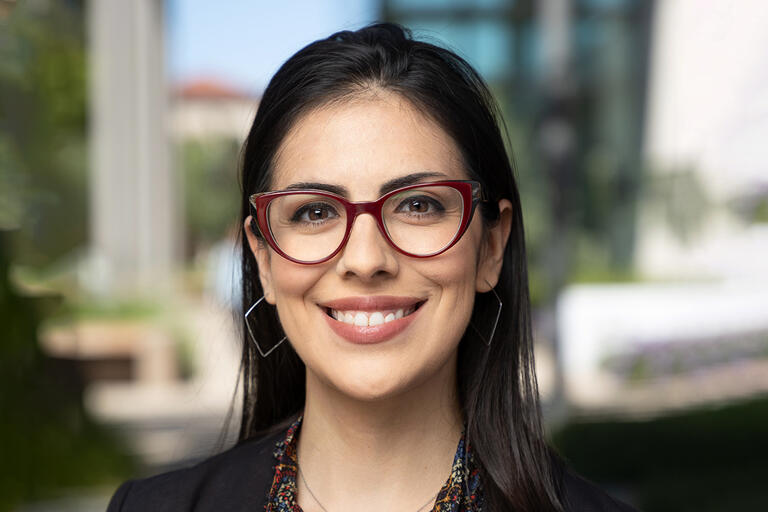 A portrait of Karina Gutiérrez. She has long black hair and brown eyes. She is wearing dark red glasses and is smiling.