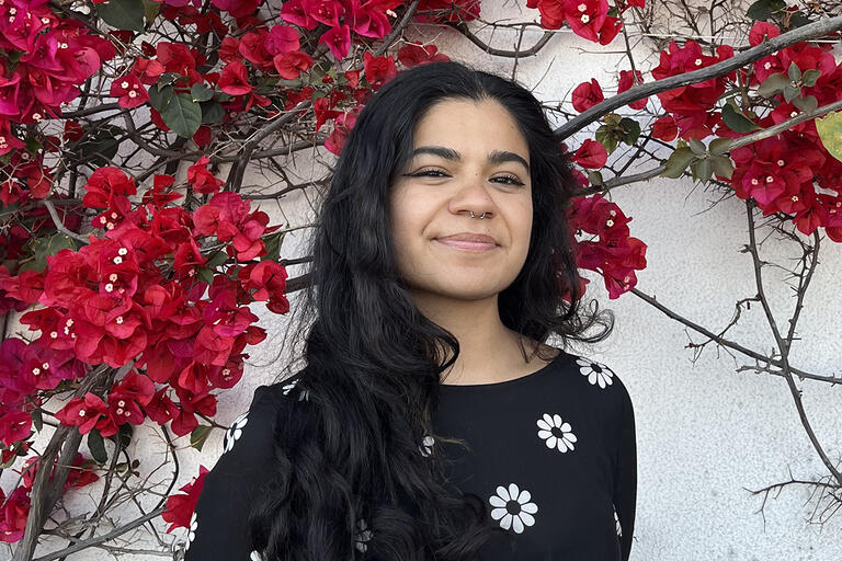 Sabrina wears a black shirt with a white flower pattern. She stands in front of a gray wall covered in red bougainvillea flowers.