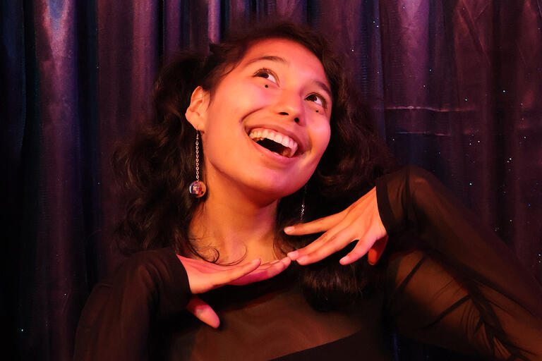 Daynna has dark, shoulder-length hair and is wearing a black shirt and small disco-ball earrings. They are holding their hands under their chin while smiling broadly and looking upward.