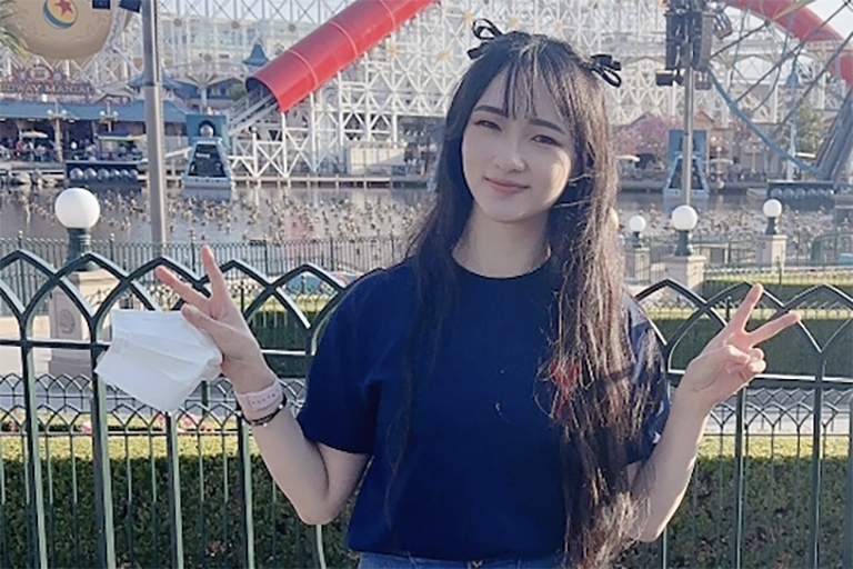 Katherine stands near a pond with a rollercoaster in the background. She looks straight ahead and smiles while holding both hands up in "peace signs."