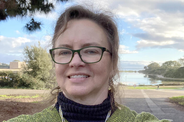 A portrait of Denise Umland. She is smiling and she is wearing a green sweater over a black turtleneck shirt. She is also wearing glasses with dark green frames.