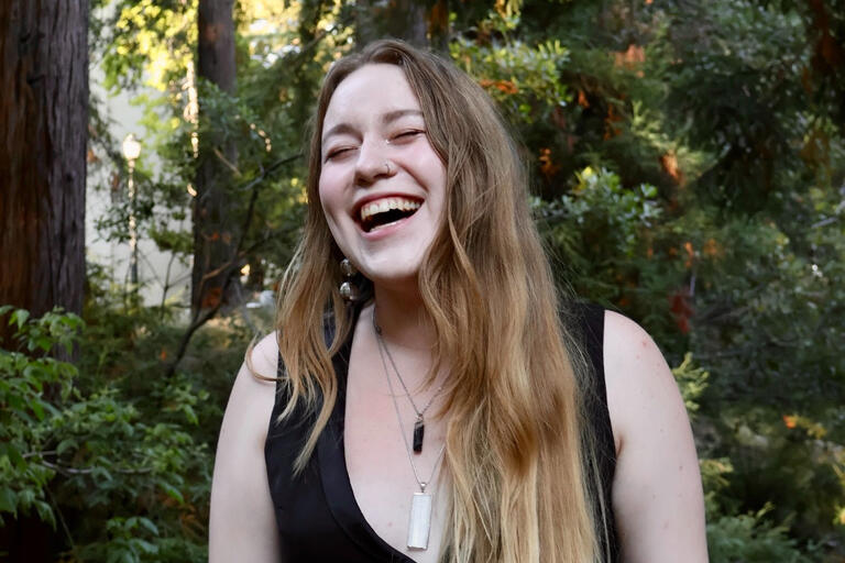 Kira is laughing with her eyes closed while standing in a grove of redwood trees. She is wearing a black blouse with white and black pendant necklaces.