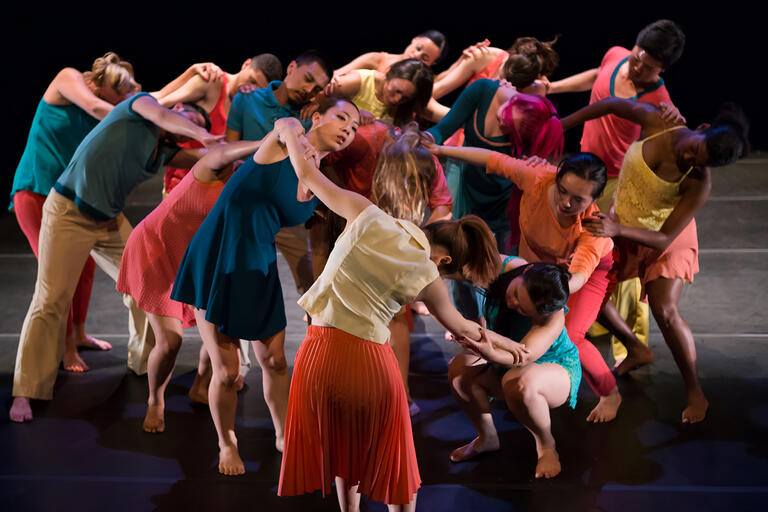 Dressed in vibrant blue, gold, and coral costumes, several dancers interlock arms in a round cluster and sway from side to side.