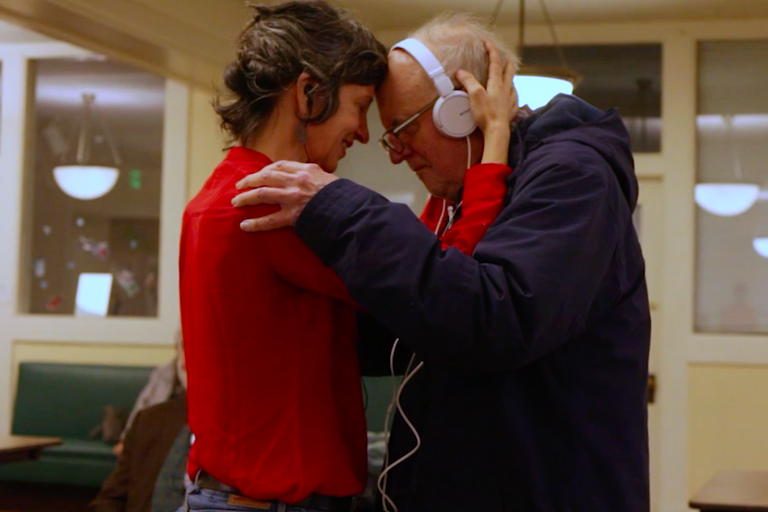 Woman in red shirt embracing elder man in blue sweatshirt wearing headphones