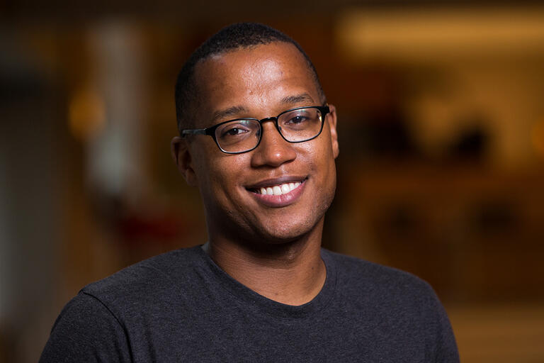 A portrait of Branden Jacobs-Jenkins. He smiles while wearing black glasses and a gray t-shirt.