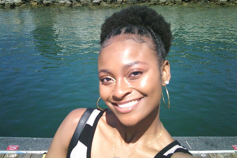 A person smiling in front of a body of water with a rocky bank in the background.
