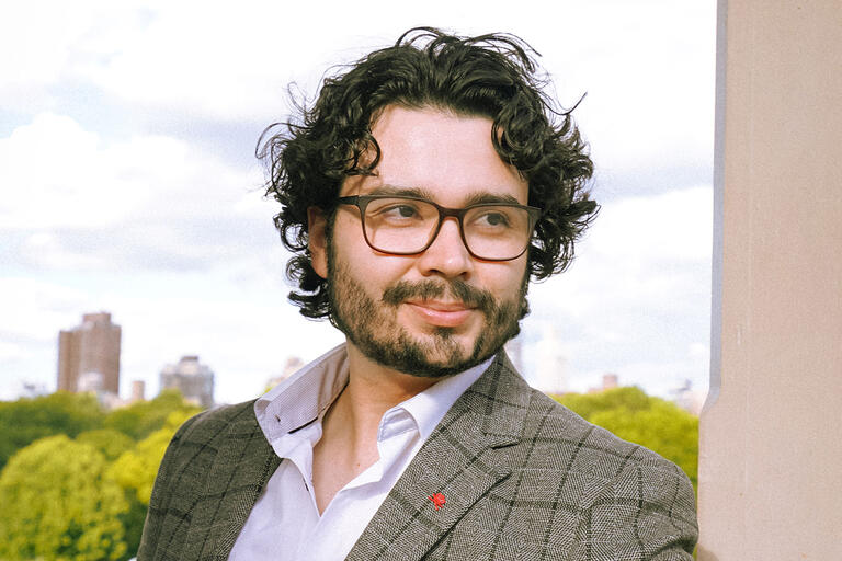 A portrait of Drew Woodson wearing dark-framed glasses, a white collared shirt, and a grey blazer. He has black wavy hair and a beard.