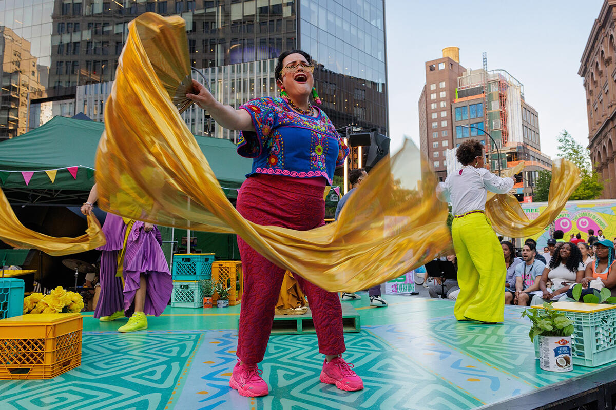 An actor in a vibrant pink and purple costume with floral motifs waves a gold silk scarf while standing on an outdoor stage in New York City.