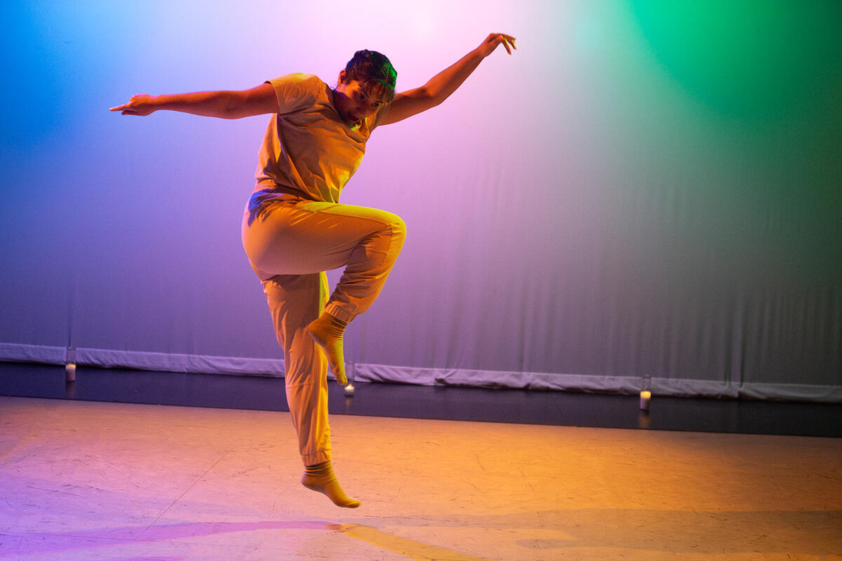 A dancer jumps in the air with one knee raised and her arms extended to either side of her body. She is illuminated in yellow light against a blue, purple, and green background.