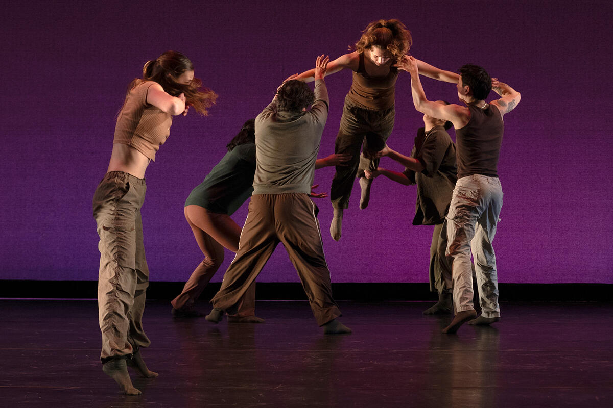 A dancer is lifted in the air by four other dancers. They are all dressed in shades of brown and green, and they are dancing against a purple background.