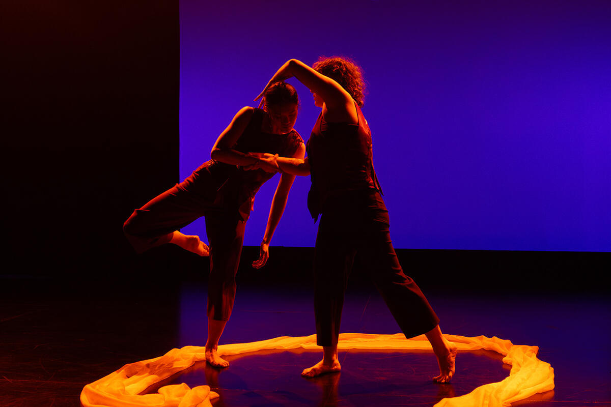 A sheer piece of fabric forms a circle on the floor around two dancers. The dancers are illuminated with golden light against a blue background.