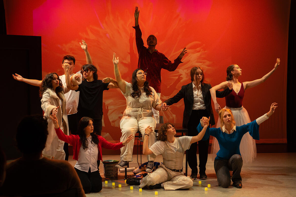 10 actors stand in front of a red-orange background with an image of a flower. Each actor looks upward and gestures with their hands in the air.