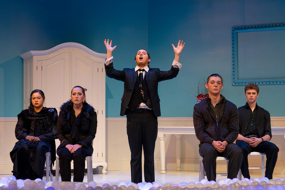 A scene from the play "Wintertime" in which four people dressed in black funeral clothes sit solemnly and stare straight ahead at the audience. Another person standing in the center delivers a eulogy while raising their hands  over their head.