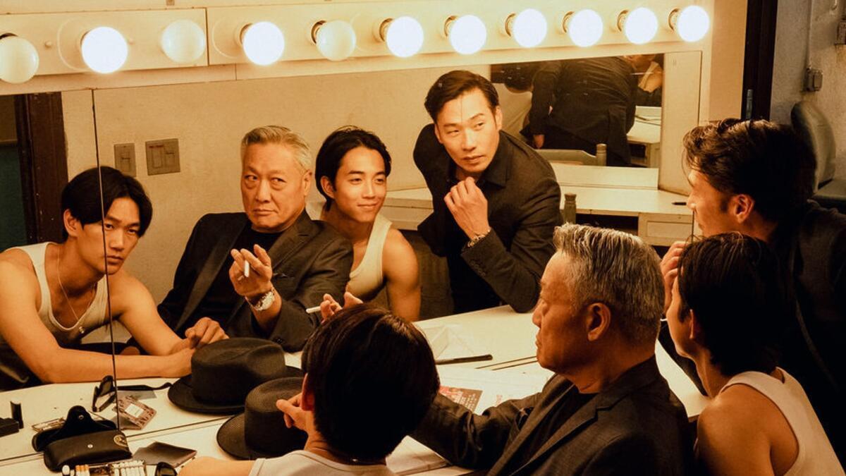 Five men gather around a brightly lit dressing room mirror, wearing a mix of suits and casual attire.