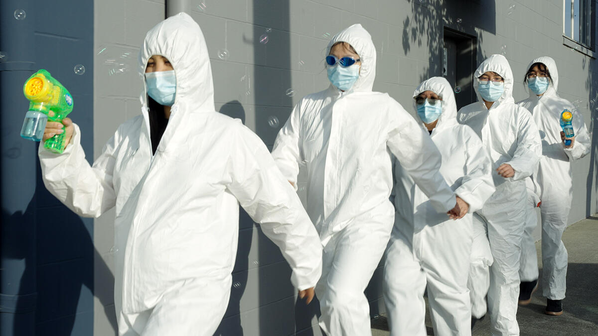 Five people wearing white biohazard suits and surgical masks walk single-file down a sidewalk. The people at the front and back of the line are carrying toy bubble makers, and soap bubbles are floating above them.
