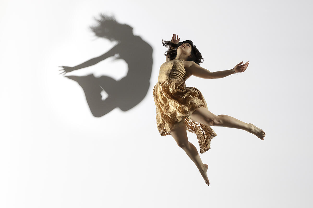 Megan Lowe wears a gold dress and leaps in the air in front of a white background. Her shadow is cast against the background in stark contrast.