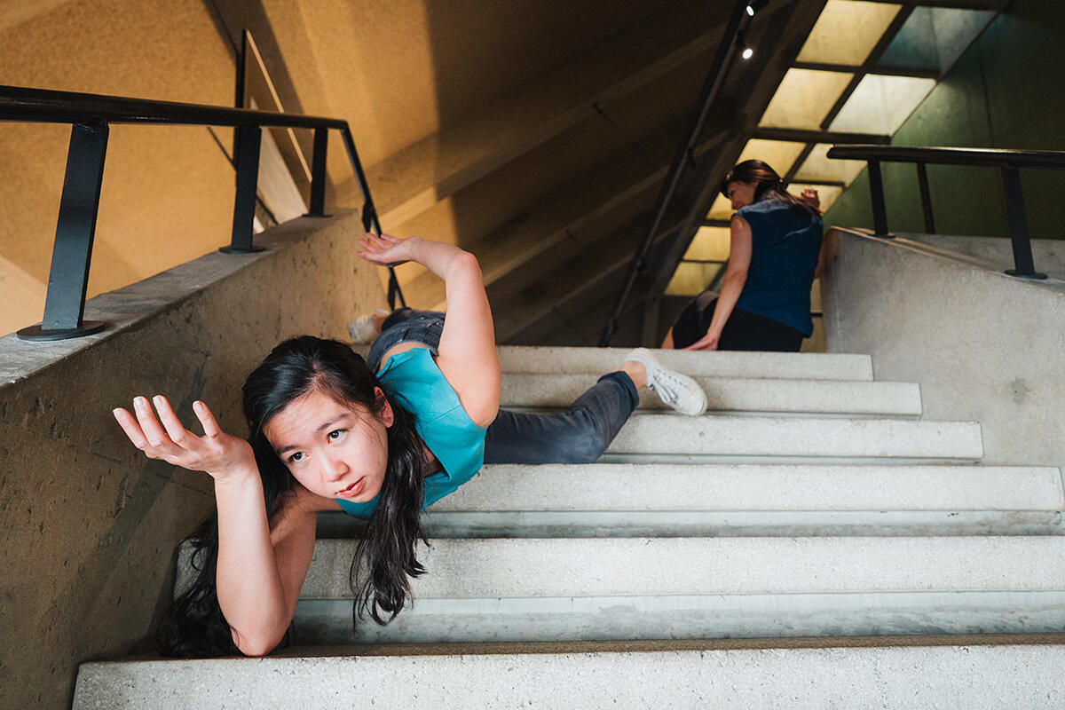 A dancer dressed in a blue shirt and jeans lies face down at a steep angle on concrete stairs. They hold their right hand in front of their face and their right arm behind their back.
