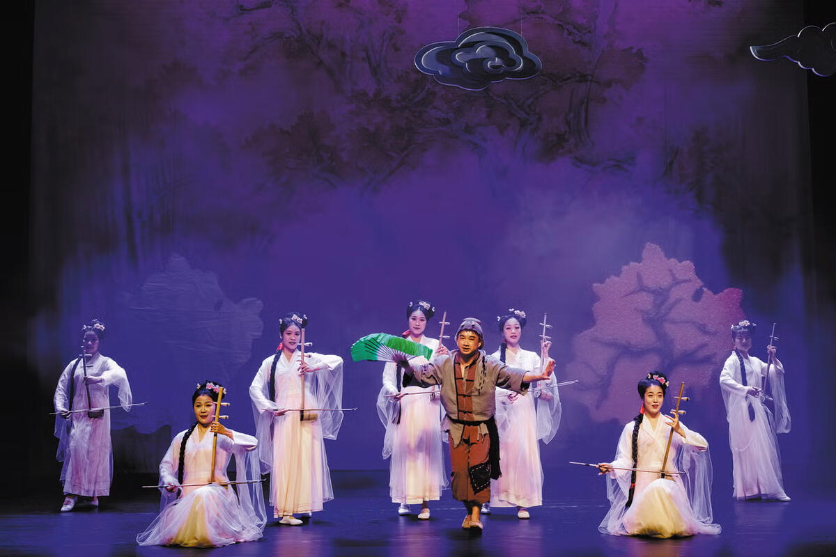 Eight performers in traditional Chinese clothing stand on a stage in front of a purple background with trees and leaves.
