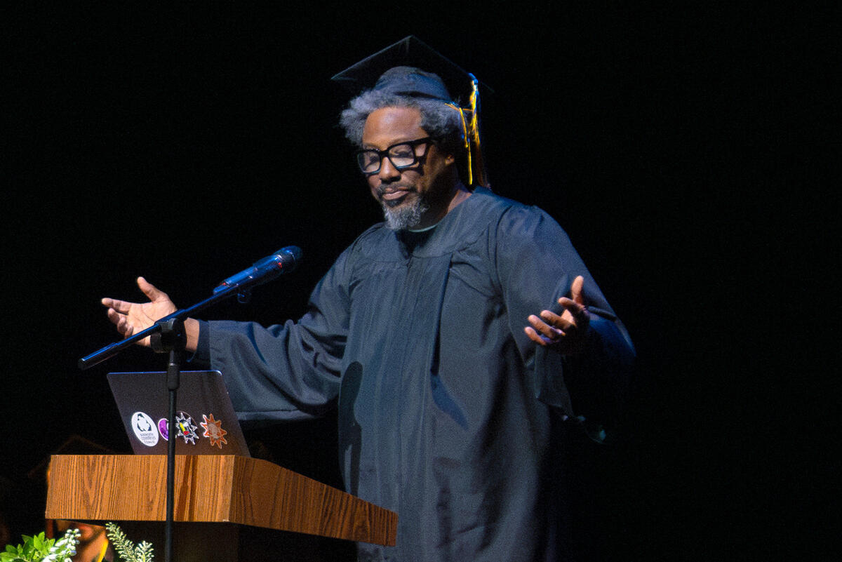 Comedian W. Kamau Bell wears a black graduation cap and gown while giving a speech at the 2024 TDPS commencement ceremony.