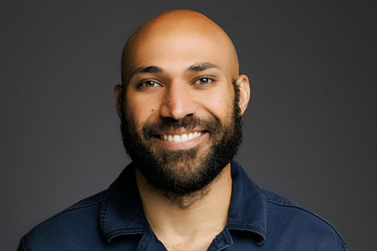 Zachary stands in front of a gray background. He is wearing a dark blue shirt and is looking forward while smiling.