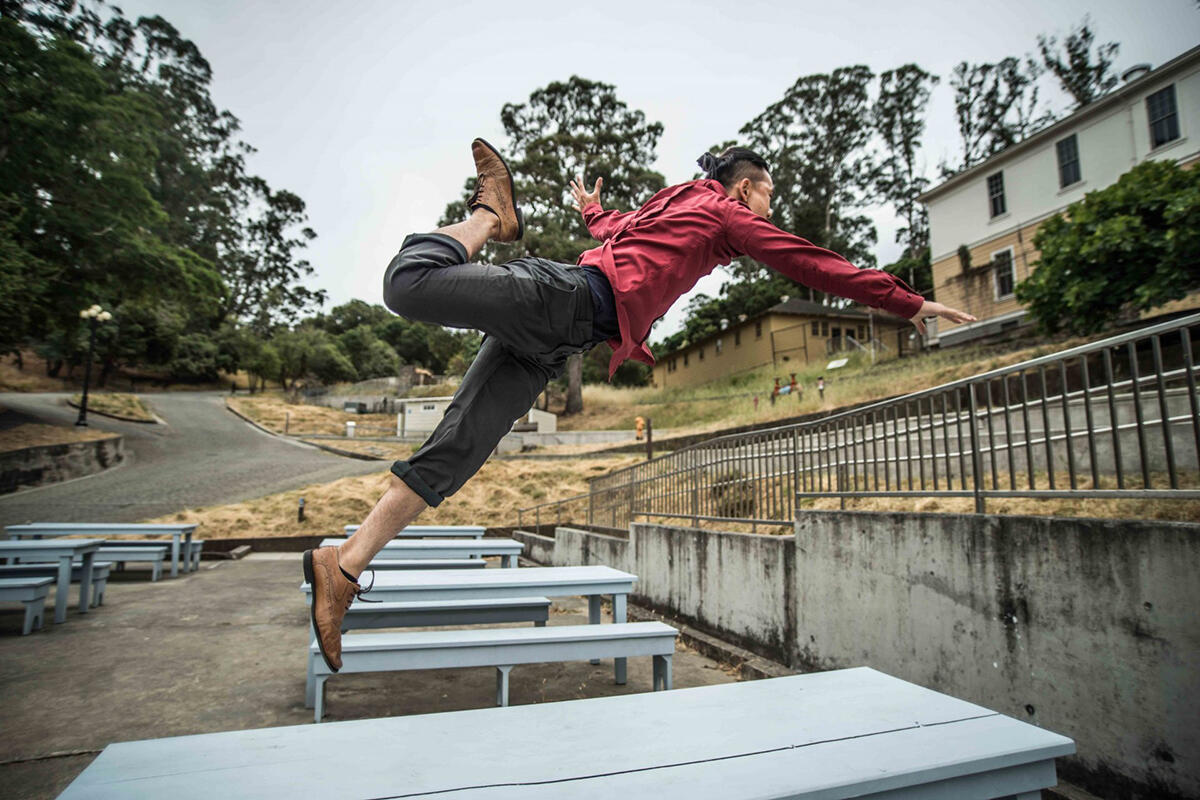 Dancer Hien Huyn wears gray pants, a red shirt, and brown Oxford shoes. He leaps several feet in the air from a gray table. In the background, we see more tables, the brown and tan barracks, and tall eucalyptus trees at the Angel Island detention center.
