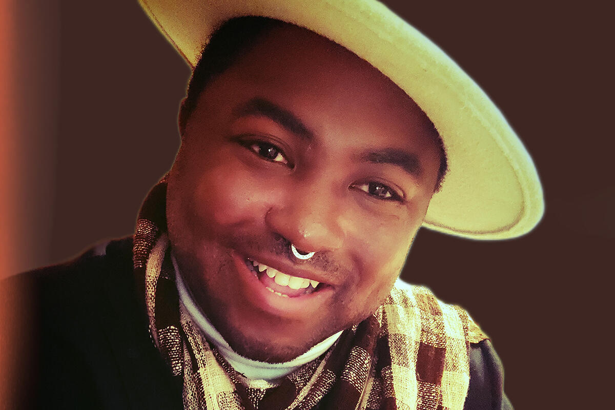 AeJay Mitchell, Graduate Student Aejay wears a wide brimmed hat, a checkered scarf, and a dark jacket. They're smiling while looking forward.